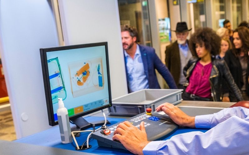 Photo of a computer showing scanned luggage in the monitor in an airport with some people in the area