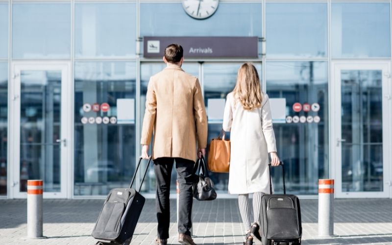 Photo of a man and woman pulling their carry on luggage heading to a building